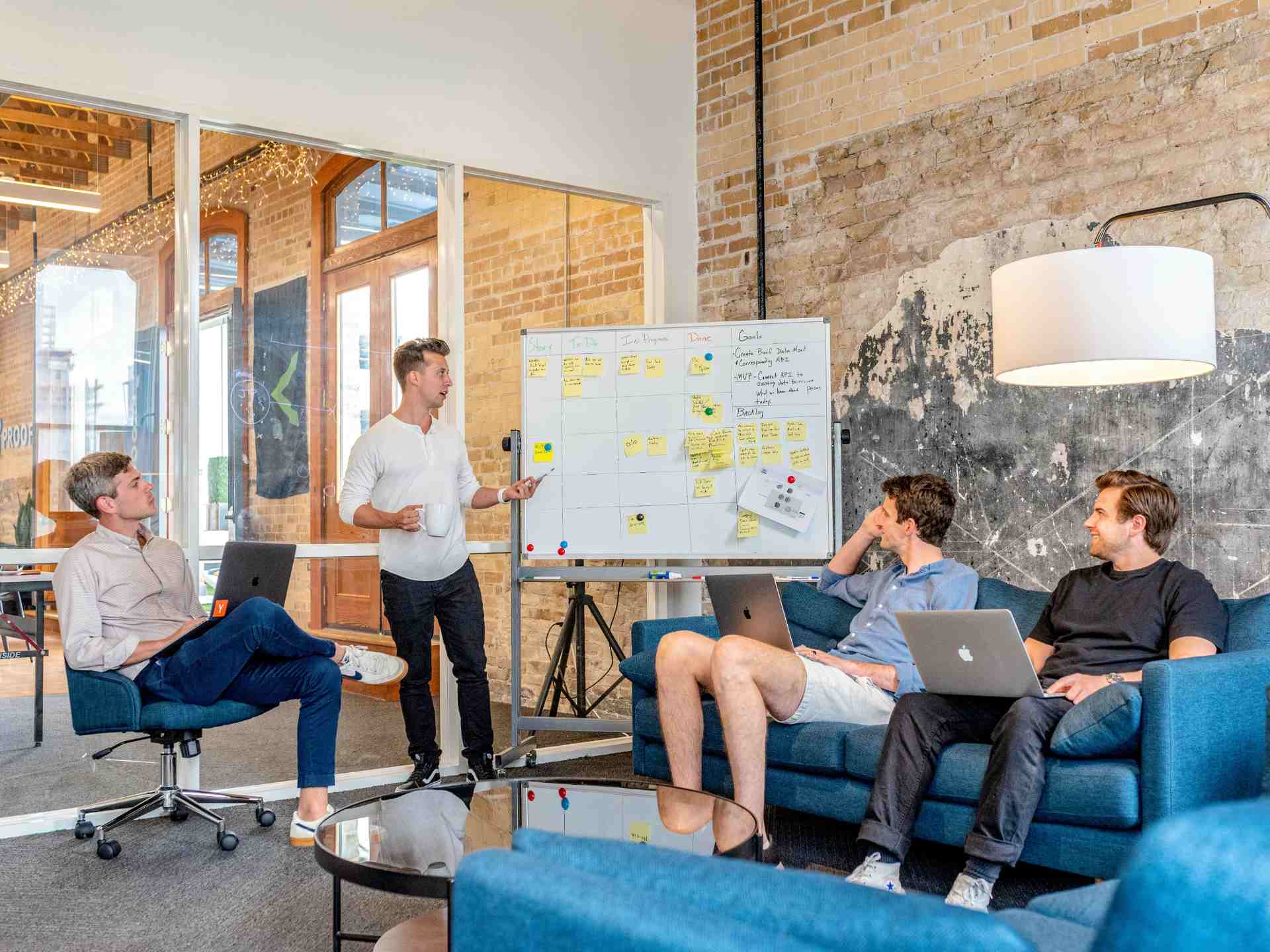 3 young professionals sitting on couches and working on their laptops while their colleague gives a presentation